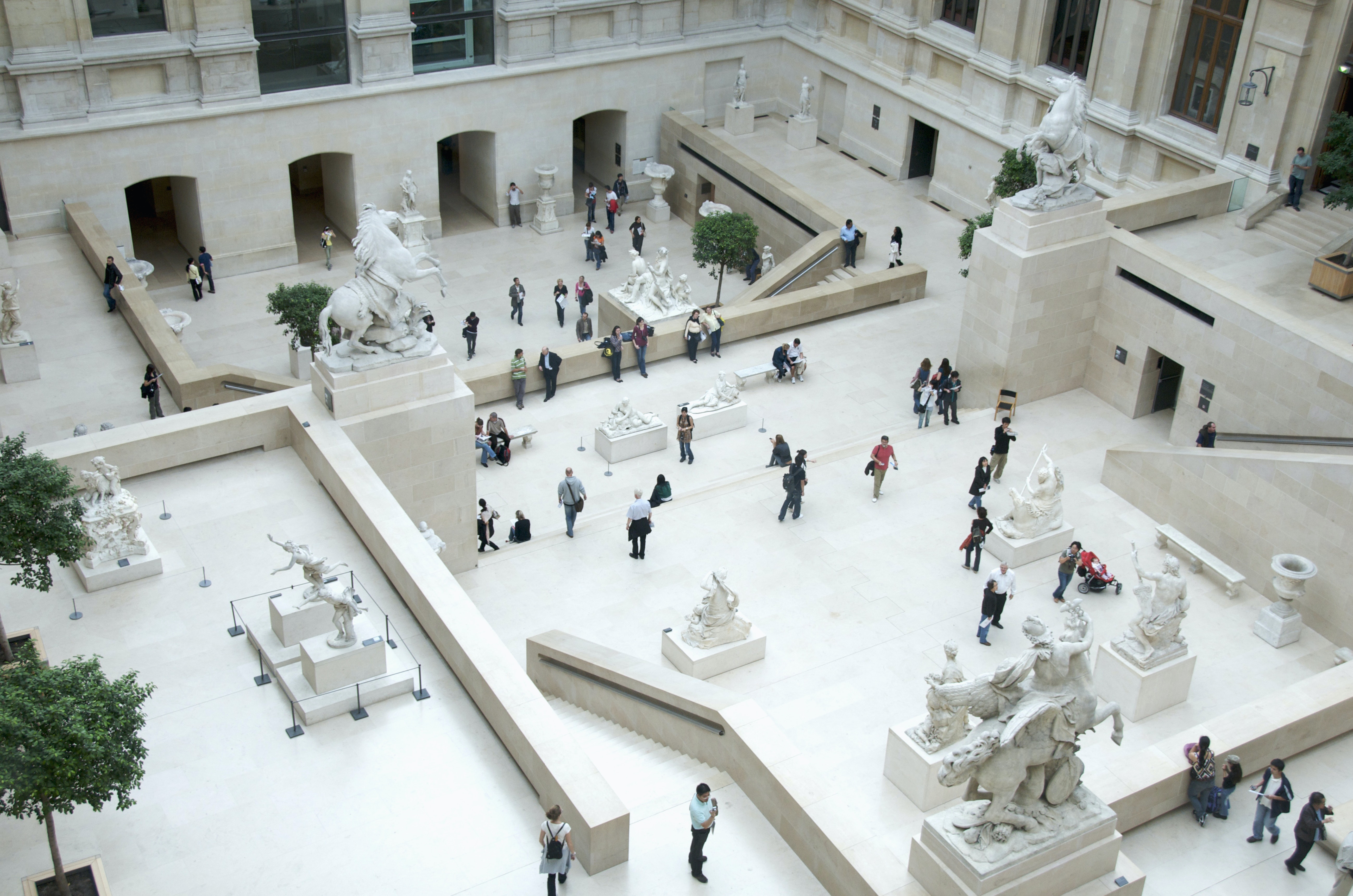 Aerial view of the Cour Marly, a courtyard in the Louvre filled with sculptures from the 17th and 18th centuries. The white courtyard is filled with visitors looking at the exhibits. 