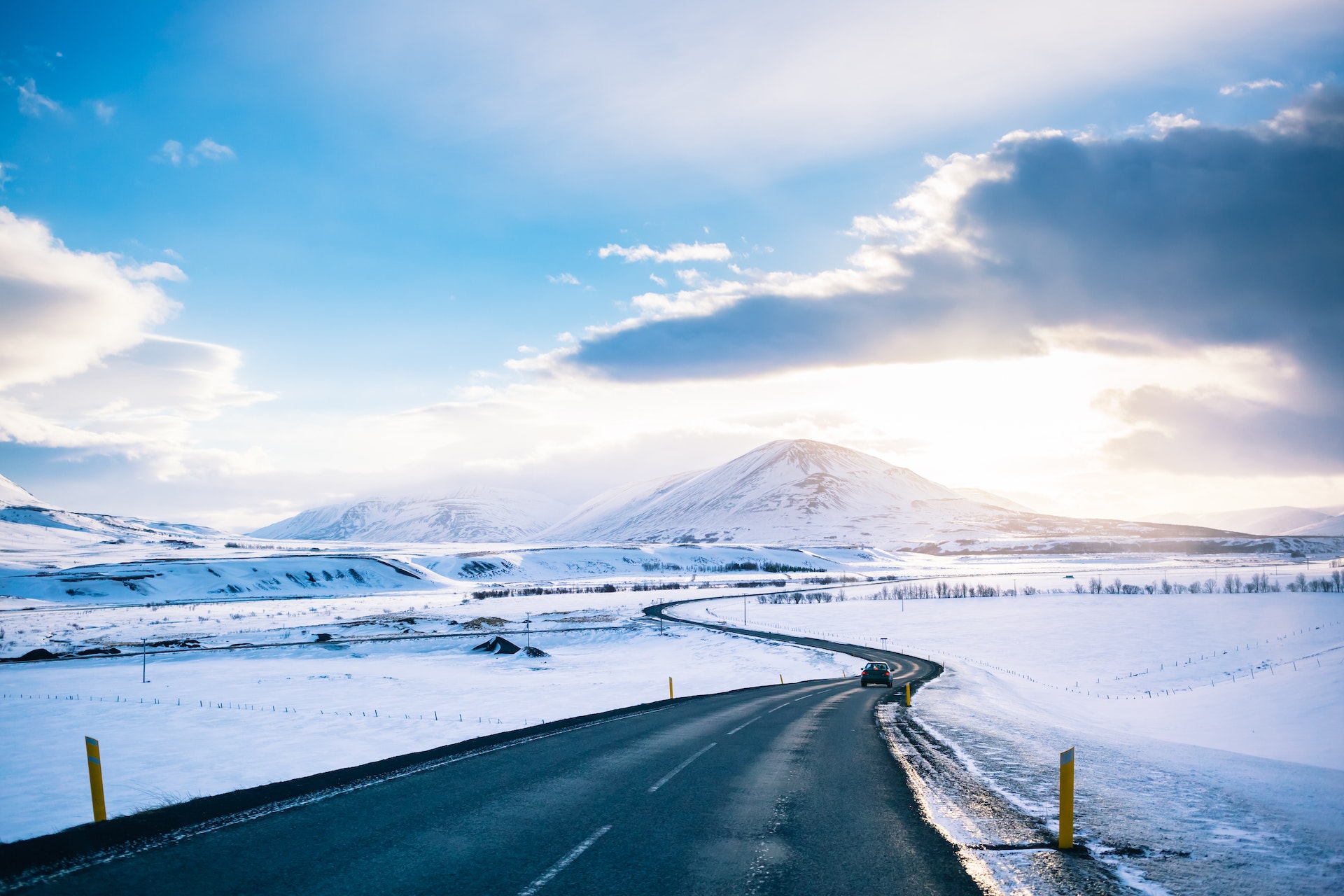 A car drives on a winter road near Akureyri, Iceland. The black road is surrounded by miles of snowy terrain. Mountains are visible in the distance.