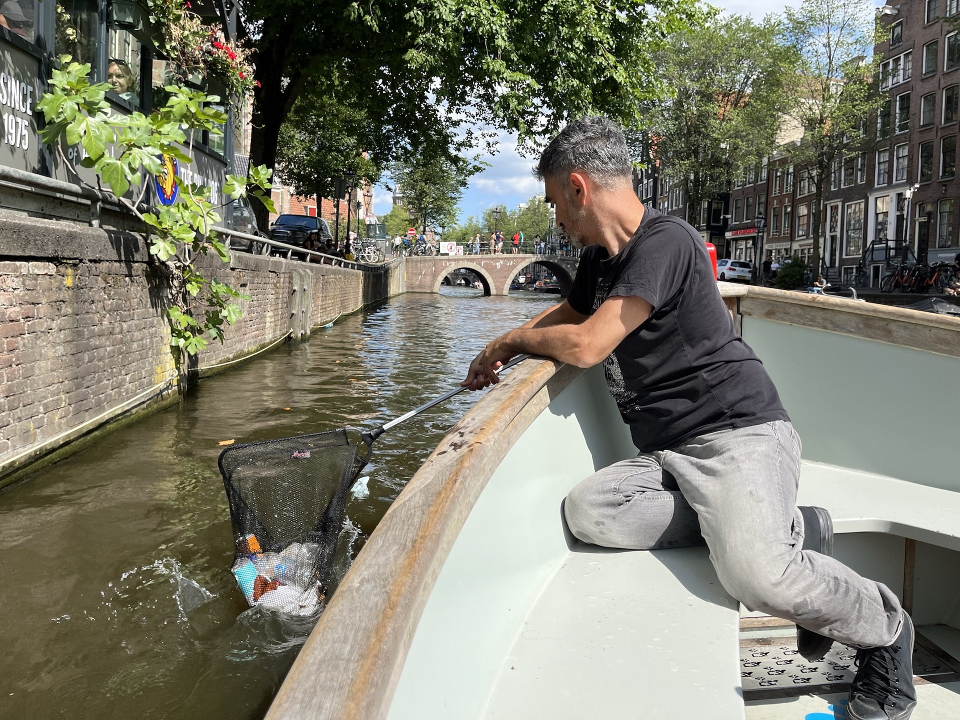 A man leans over the edge of a fishing boat to grab trash from the canal with his net